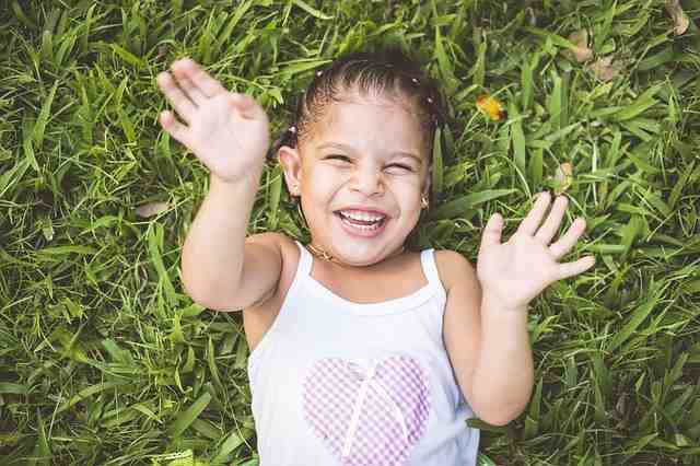 Young girl laying in the grass with a white tank top on holding her hands up and laughing, image used for Ari Monkarsh blog that children are the future and we should teach them to be philanthropic