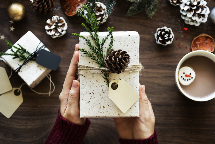 Nicely wrapped box in white wrapping paper tied with twine and a pine cone and spruce sprig decorating it as woman holds it over a table with pinecones, hot chocolate, and a notebook on a table, image used for ari Monkarsh post about giving ethical holiday gifts